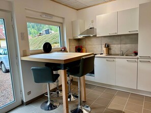 Une cuisine moderne avec un îlot central en bois, deux tabourets de bar, une fenêtre donnant sur une maison et des arbres, et des meubles de rangement blancs.