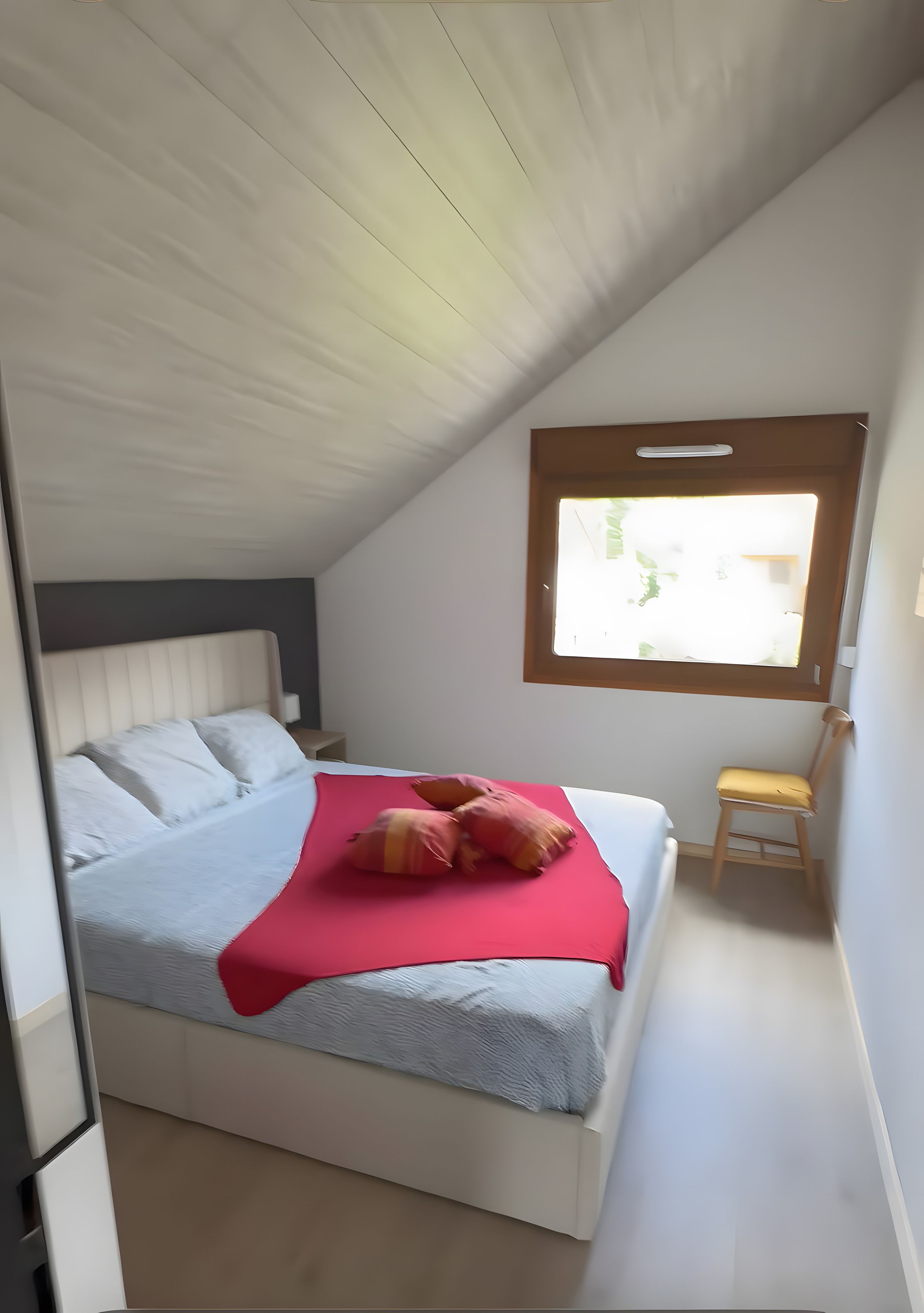 Ein Schlafzimmer mit schräg verlaufender Decke, einem Bett mit weißem Kopfteil, einer roten Decke und einem kleinen Holzstuhl.