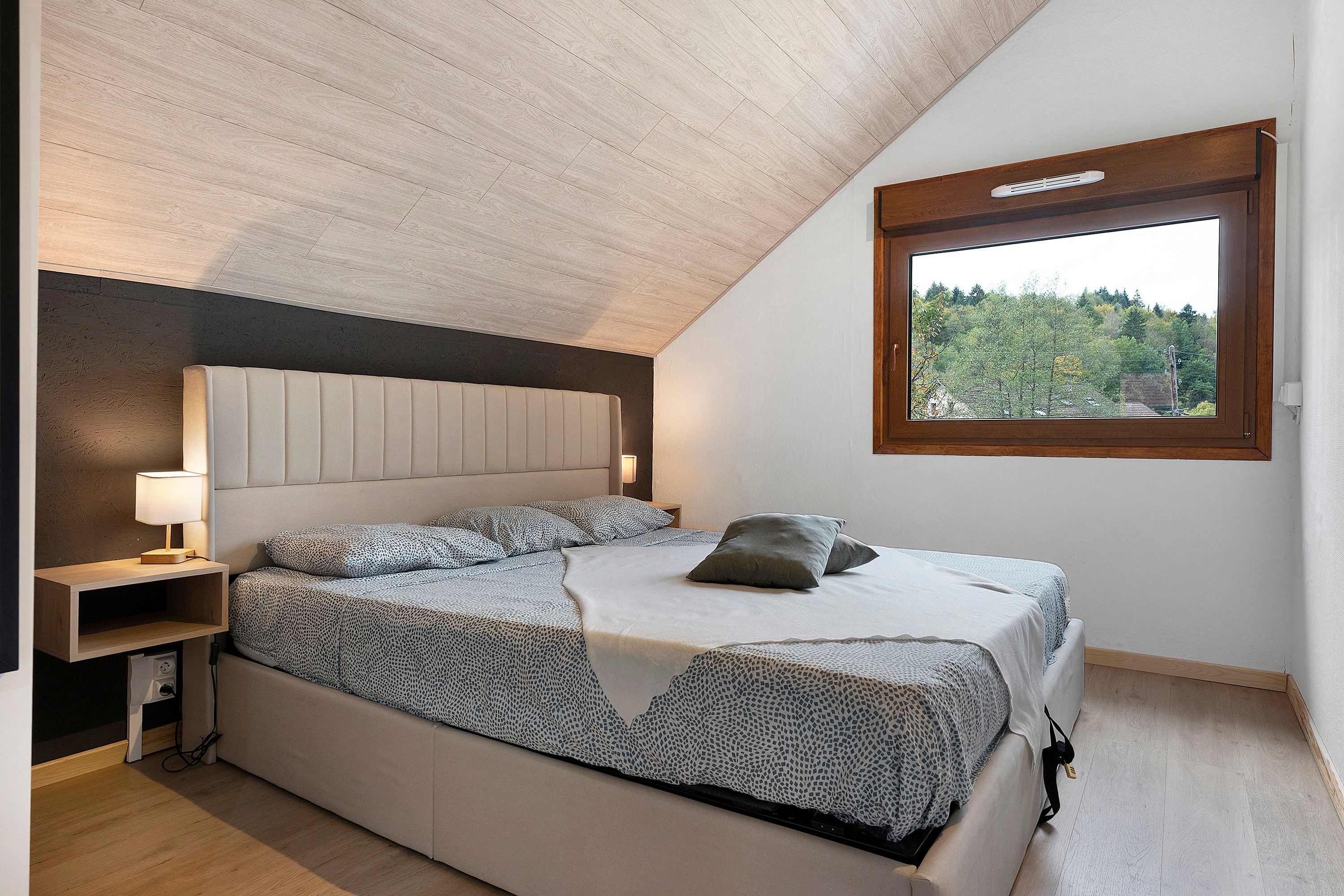 A bedroom with a sloped ceiling, a bed with patterned bedding, a bedside lamp, and a window with a view of trees.