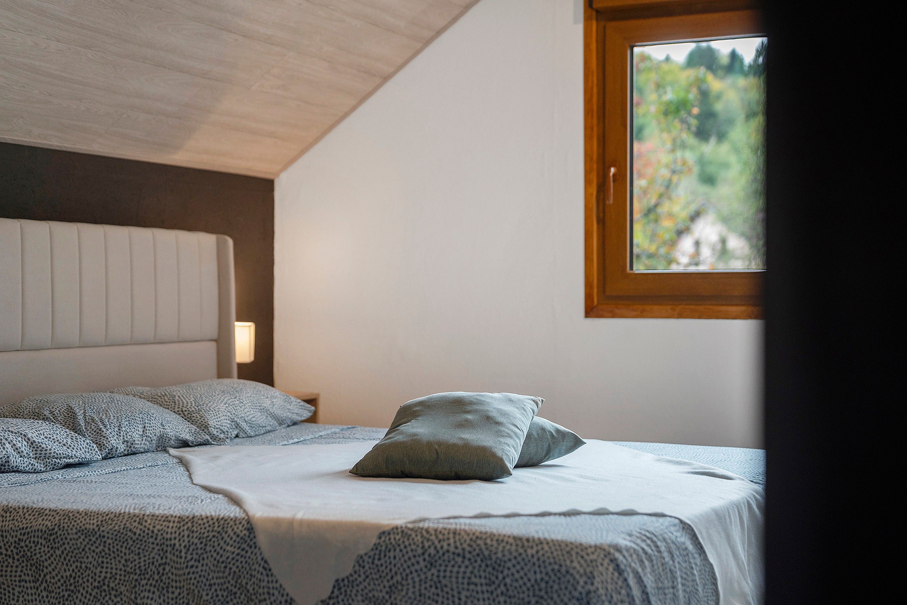 A bedroom with a bed, pillows, a headboard, and a window with a view of greenery.