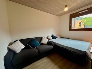 A modern bedroom with a black sofa, a bed, and a window with a view of a house. - Chalet 'Le Bonheur Vosgien' (Ventron)