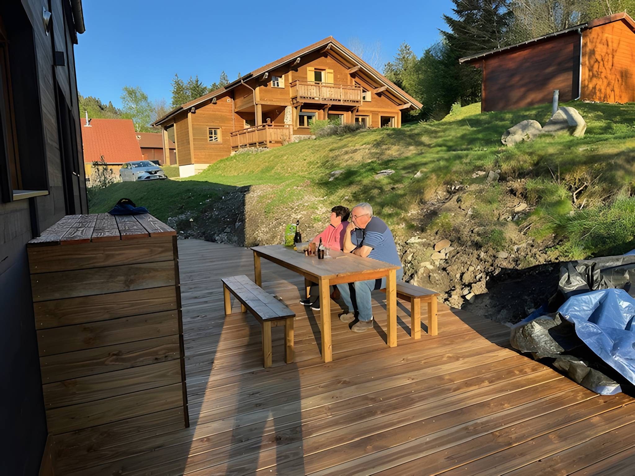 A wooden deck with a table and benches, overlooking a house and a grassy hill.