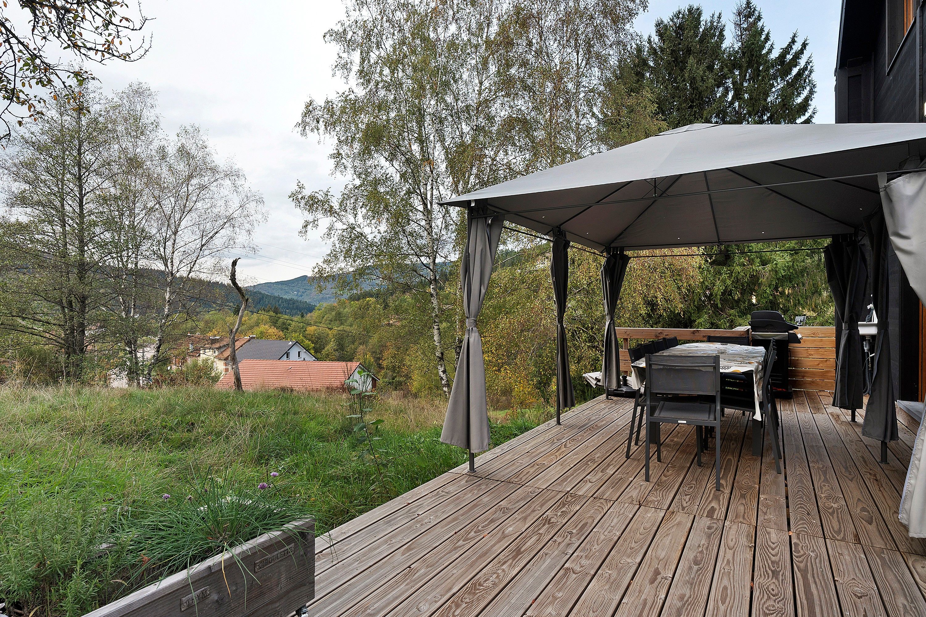 A wooden deck with a covered outdoor dining area, a grill, and a view of a house in the background.