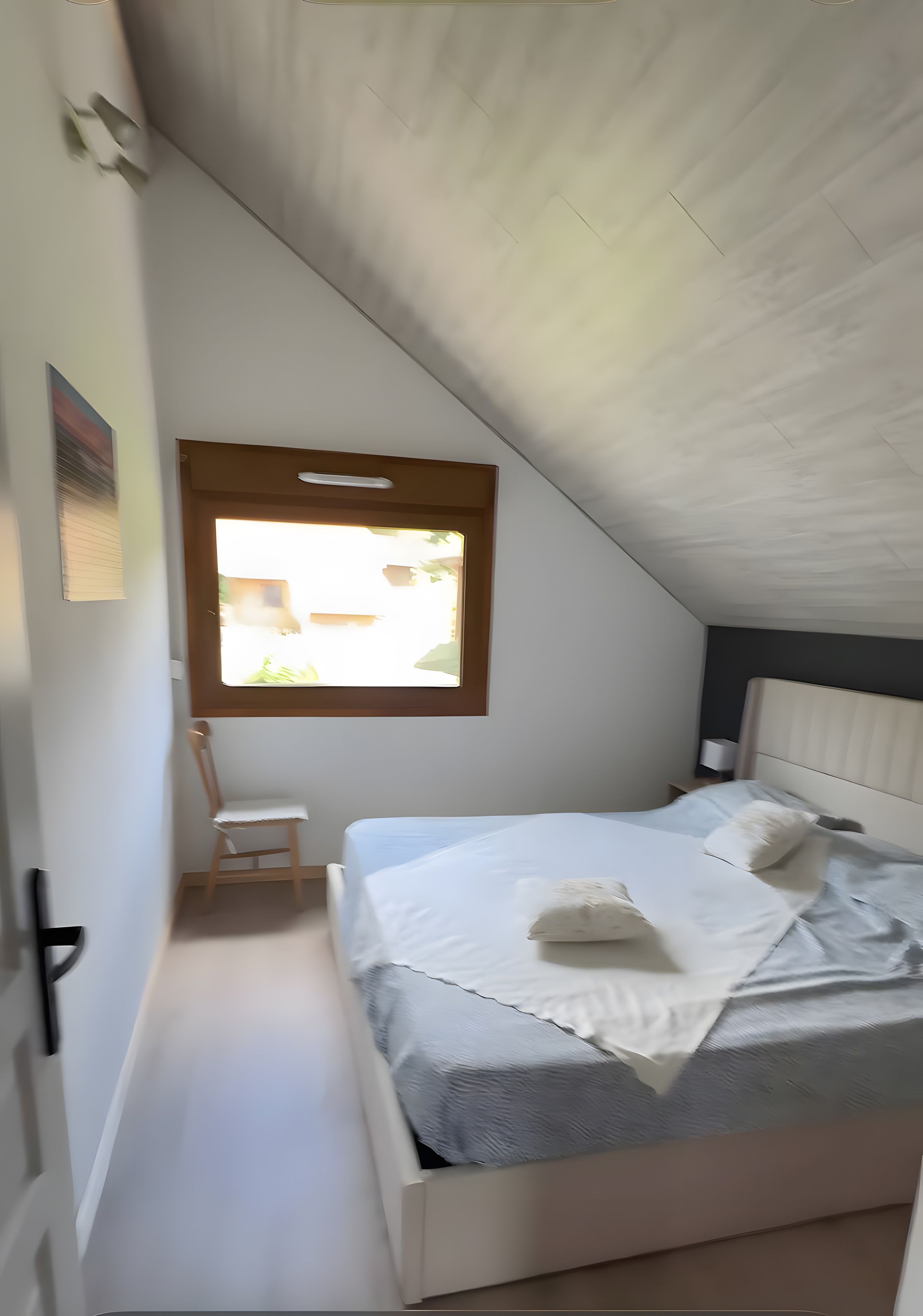 Ein Schlafzimmer mit schräg angesetzter Decke, einem Bett mit weißer Bettwäsche, einem Fenster mit Blick und einem Sessel.