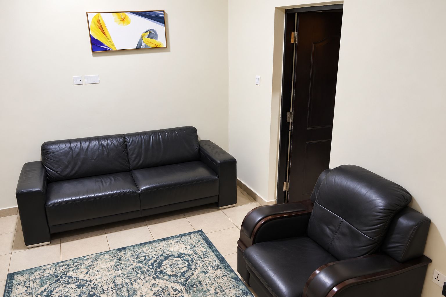 Una sala de estar con un sofá de cuero negro, un sillón a juego, un tapete estampado y un cuadro en la pared.