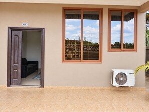 Een gebouwde ingang met een deur waarop 'FLAT 3' staat, een raam met uitzicht op een hek en bomen, en een airconditioner aan de rechterkant.