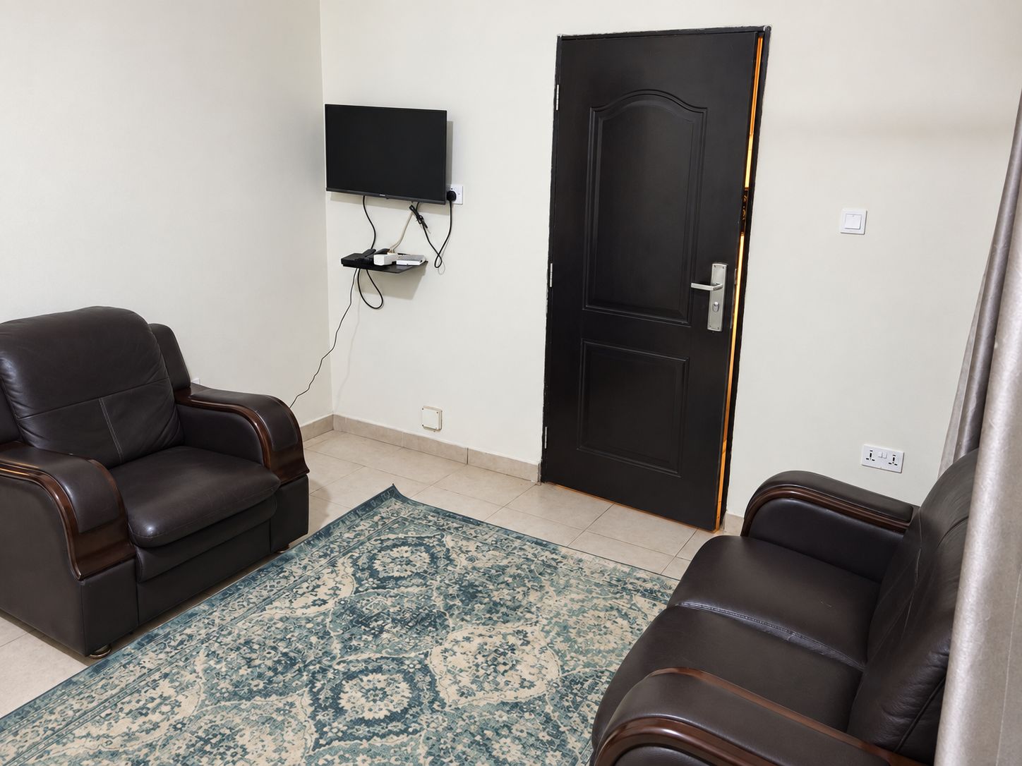 Une pièce avec deux canapés en cuir, un tapis à motifs, une porte noire et un téléviseur fixé au mur.