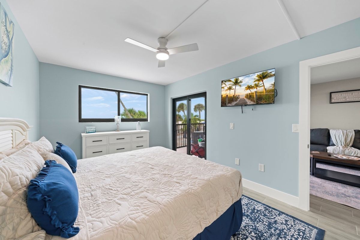 A bedroom with a bed, a ceiling fan, a window, a TV, and a door leading to a balcony.