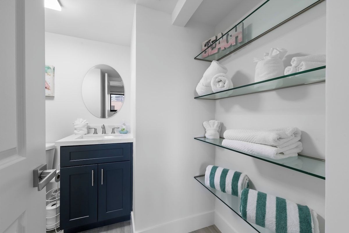 Une salle de bain moderne avec un meuble bleu foncé, un miroir rond et des étagères en verre où sont pliés des serviettes.