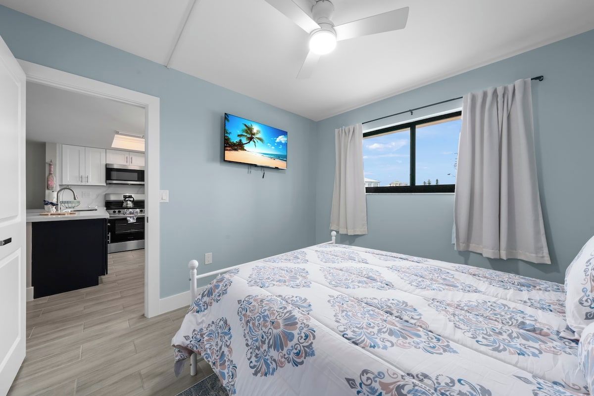 A bedroom with a bed, a ceiling fan, a window with curtains, a painting on the wall, and a view of the kitchen.