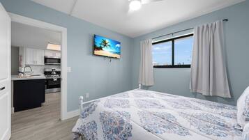 A bedroom with a bed, a ceiling fan, a window with curtains, a painting on the wall, and a view of the kitchen.