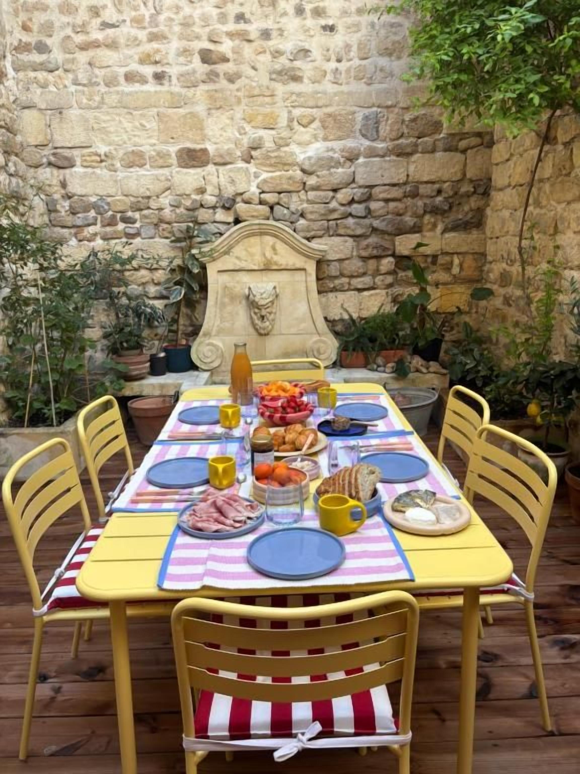 A stone wall, a table set with plates, cups, and food, surrounded by yellow chairs.