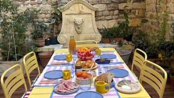 Un muro in pietra, un tavolo apparecchiato con piatti, tazze e cibo, circondato da sedie gialle.