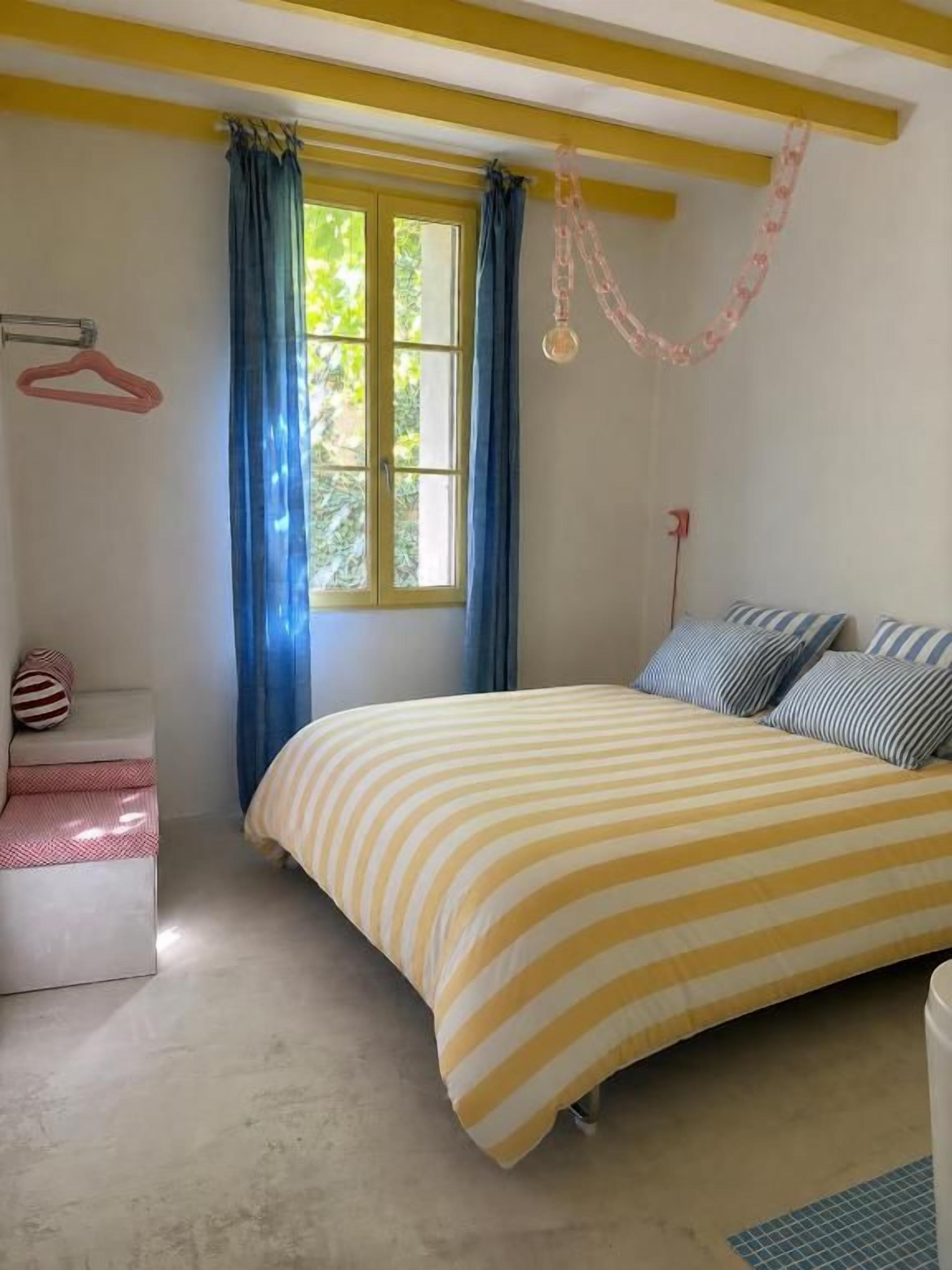 A bedroom with a bed, a window with curtains, a hanging light fixture, and a small bedside table.