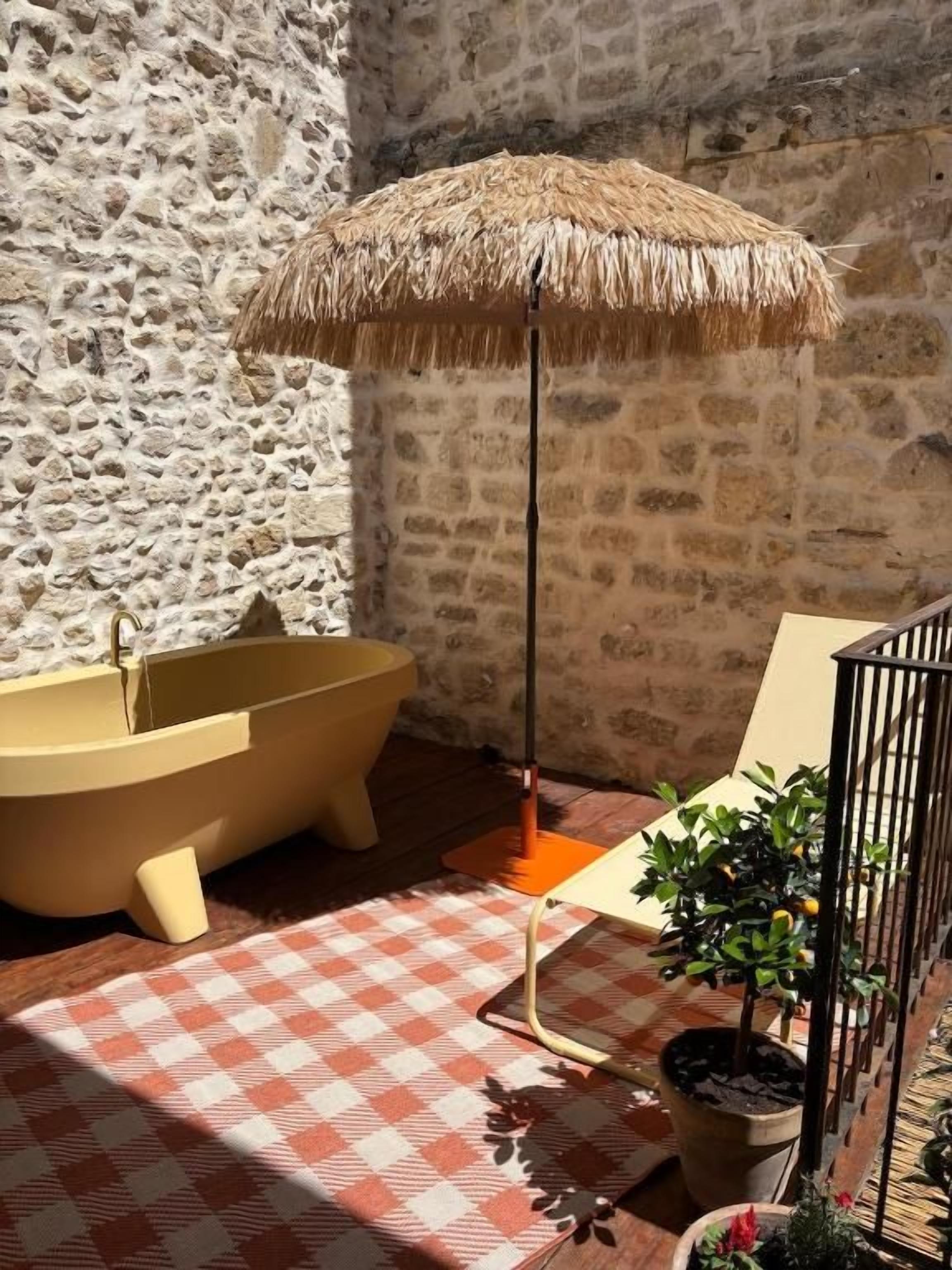 A stone wall, a bathtub, a checkered floor, a straw umbrella, and potted plants.