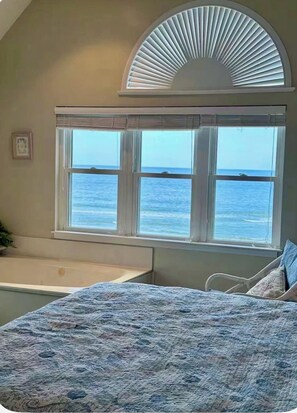 A bedroom with a large window overlooking the sea, a bathtub, and a bed with a patterned bedspread. - Cape Coddages 205 · Oceanfront 2BR Surfside Beach (Surfside Beach)