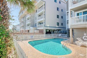 A swimming pool with a white fence, lounge chairs, and a multi-story building with balconies. - Cape Coddages 205 · Oceanfront 2BR Surfside Beach (Surfside Beach)