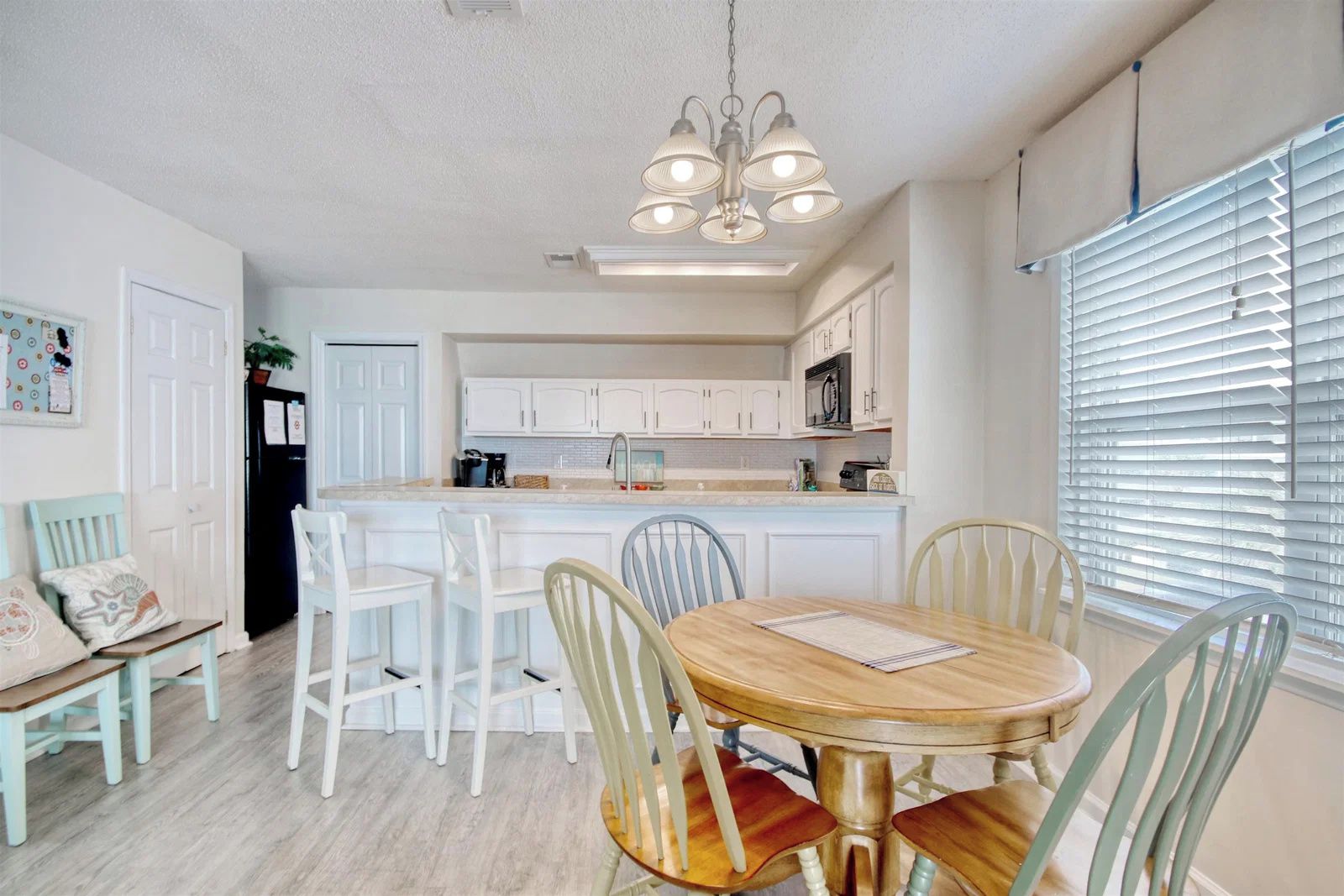 A dining area with a round table and chairs, a kitchen in the background, and a window with blinds.