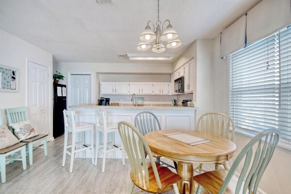 A dining area with a round table and chairs, a kitchen in the background, and a window with blinds. - Cape Coddages 205 · Oceanfront 2BR Surfside Beach (Surfside Beach)