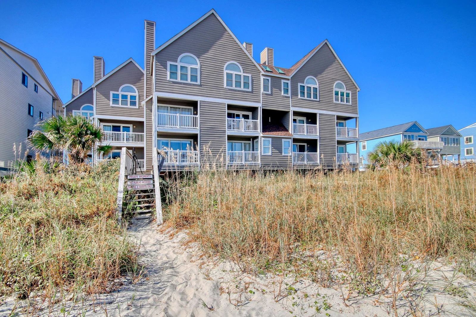 Un immeuble résidentiel à plusieurs étages situé en bord de mer, doté de balcons, d’une pancarte indiquant qu’il s’agit d’une propriété privée et d’un chemin de sable menant à l’entrée.