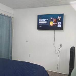 Une chambre avec un lit, un téléviseur fixé au mur qui diffuse une publicité pour un abonnement groupé à Disney+ et Hulu, et un ventilateur sur pied.