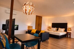 Ein modernes Hotelzimmer mit Holzboden, einer schwarzen Couch mit gelben Kissen, einem Bett mit dunklem Kopfteil und einem Essbereich mit Tisch und Stühlen.