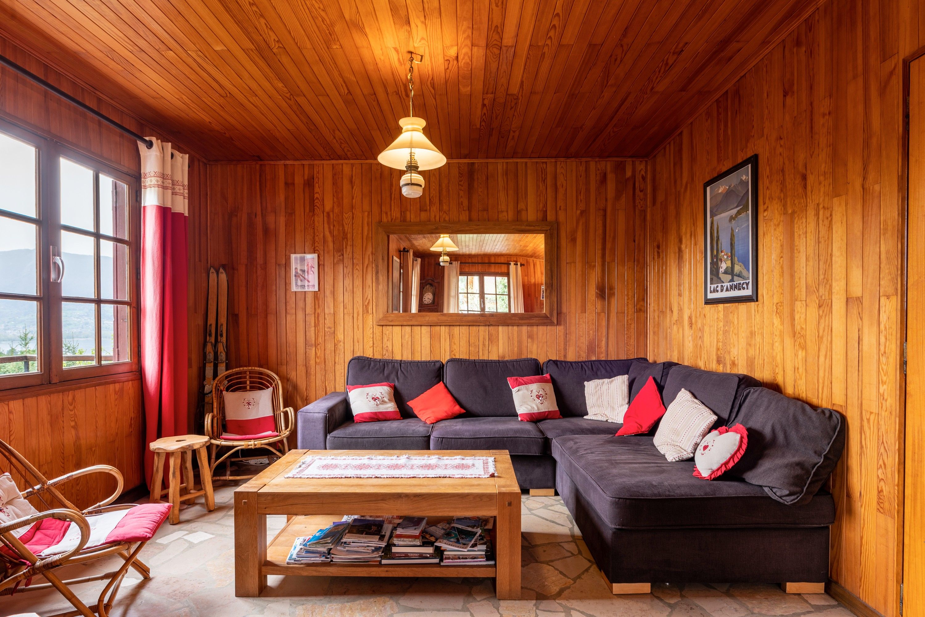 A cozy room with wooden walls, a dark sofa with red and white cushions, a wooden coffee table with books, and a wicker chair.