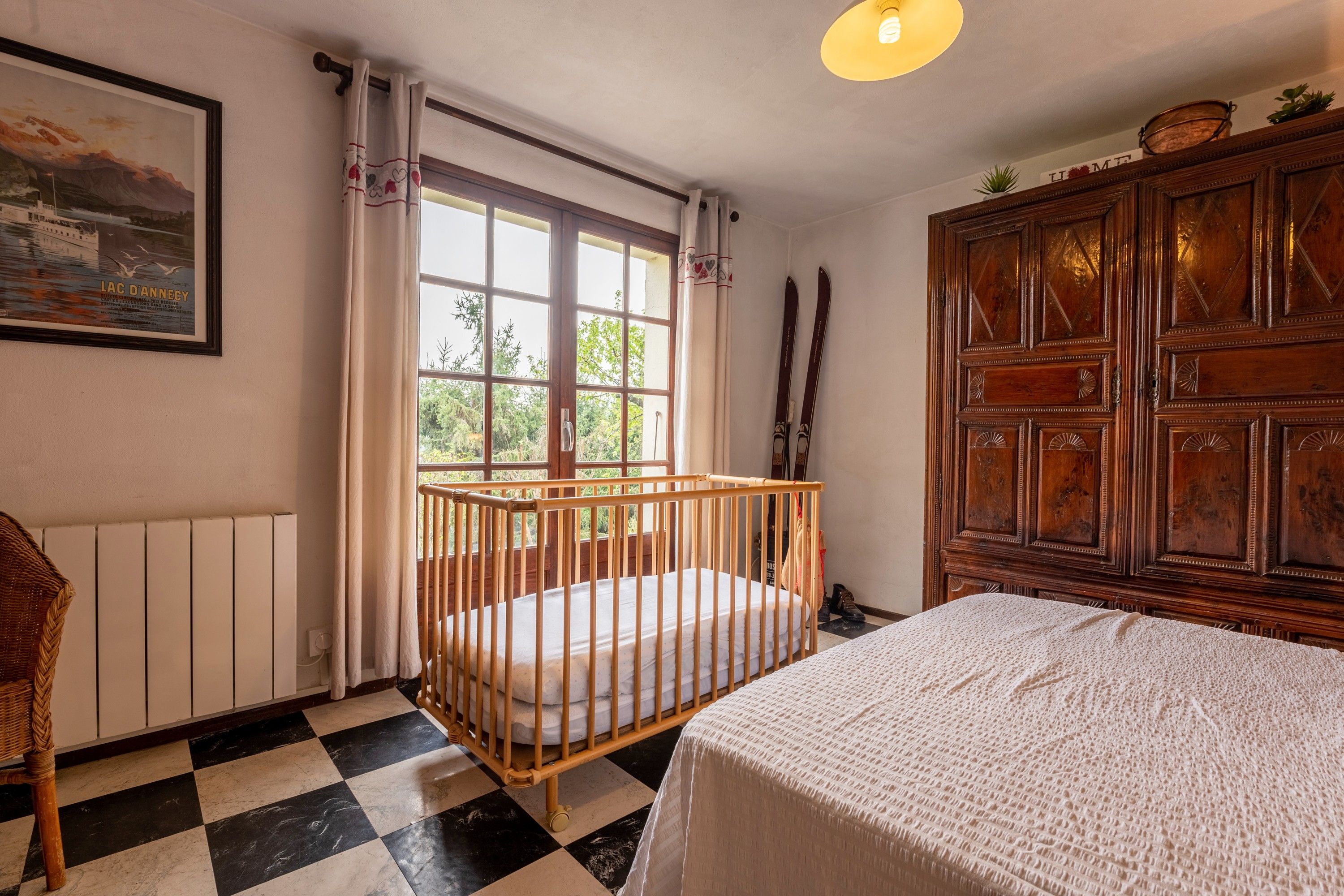Une chambre avec un berceau, un lit, une armoire en bois, une fenêtre avec des rideaux et un radiateur.