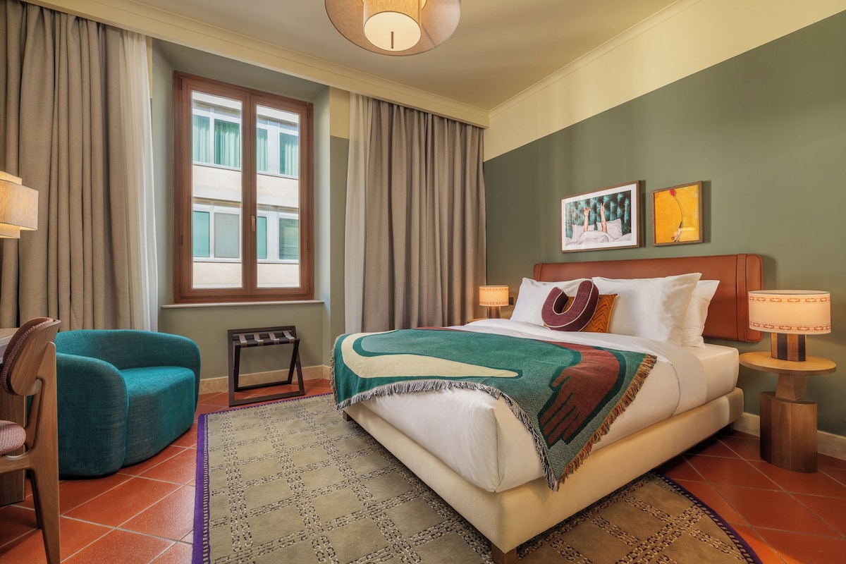 Een hotelkamer met een bed, twee stoelen, een raam, een schilderij en een lamp.