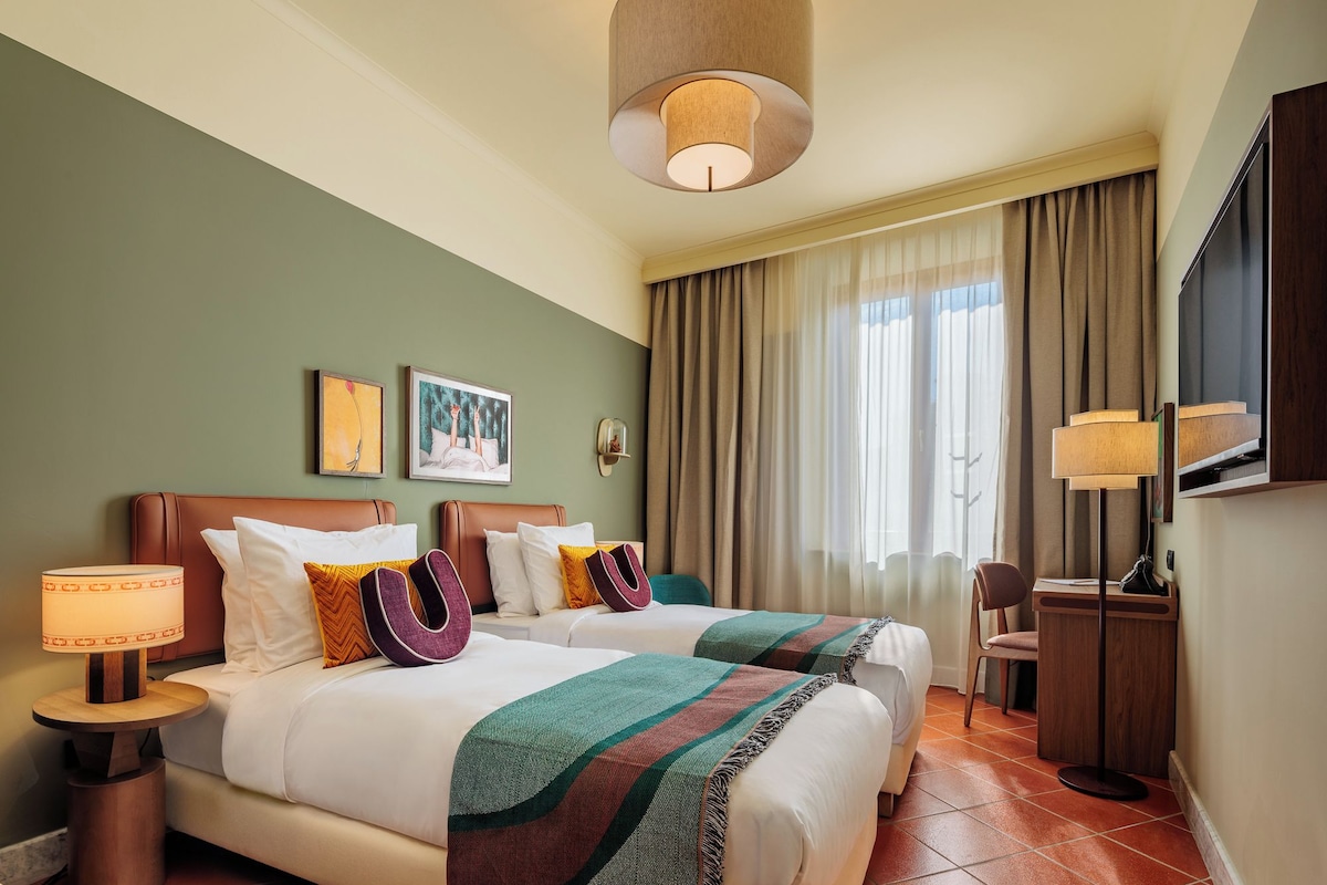 Een hotelkamer met twee bedden, een bureau, een stoel, een televisie en kunst aan de muur.