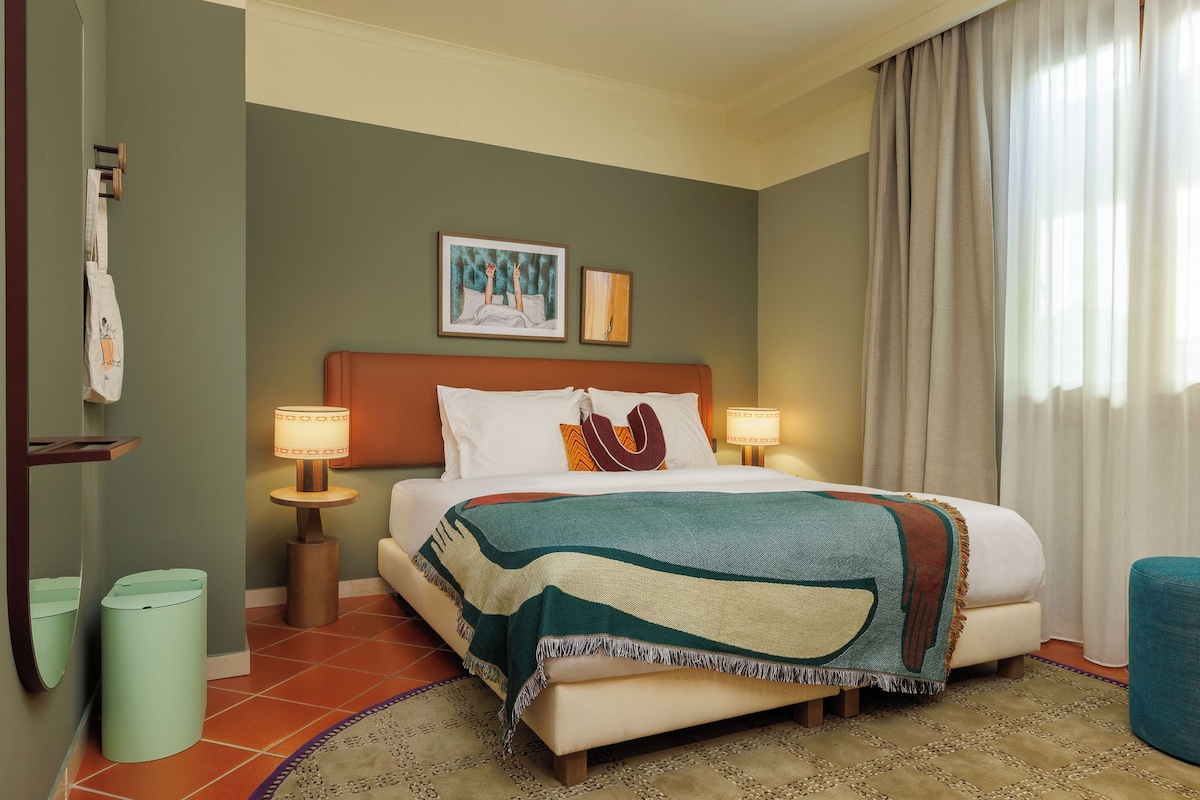 Een hotelkamer met een bed, twee bedlampen, een hoofdeinde, een groot raam met gordijnen, een wandrek voor handdoeken en een kleine prullenbak.