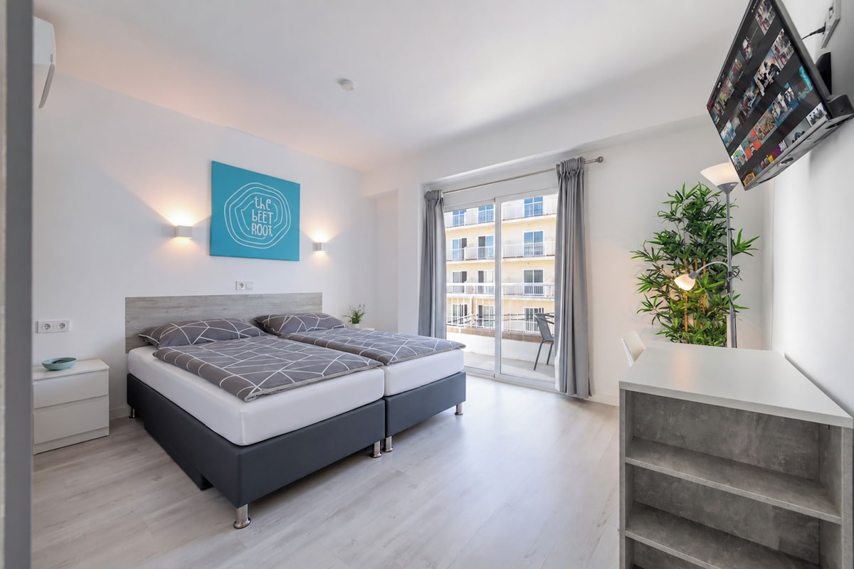 Quarto moderno com cama, TV na parede, planta e vista para a varanda externa.
