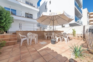 Uma área de pátio externa com cadeiras e mesas de plástico branco, um grande guarda-sol e um cachepot de pedra com um cacto.