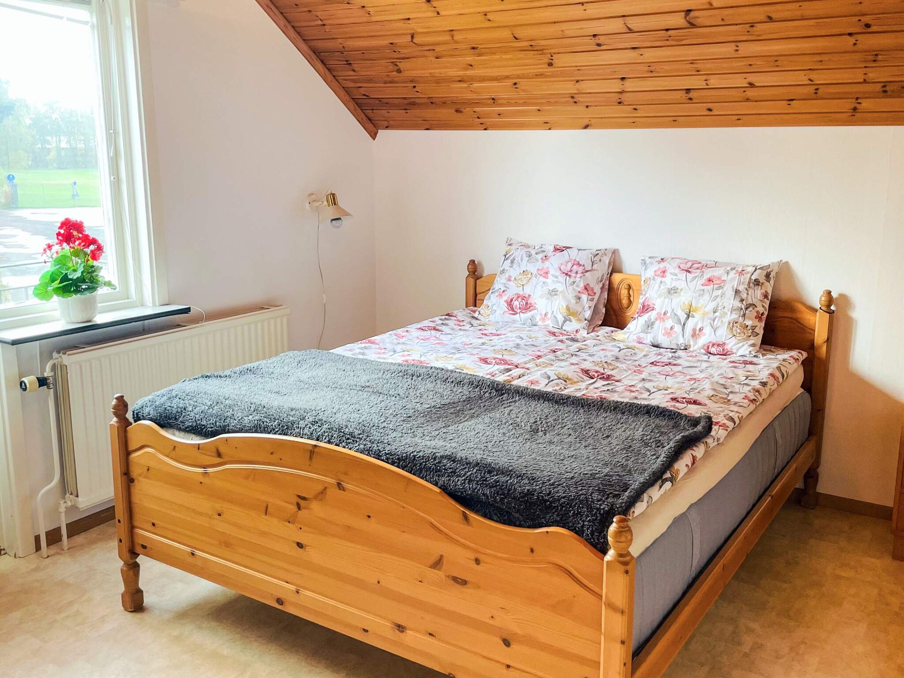 A wooden bed with a floral bedspread and pillows, a radiator, a window with a plant, and a wooden ceiling.