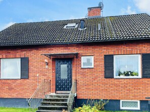 A red brick house with a black door, two windows with shutters, a satellite dish, and a small garden. - 8 Person Holiday Home in Stockaryd (Stockaryd)