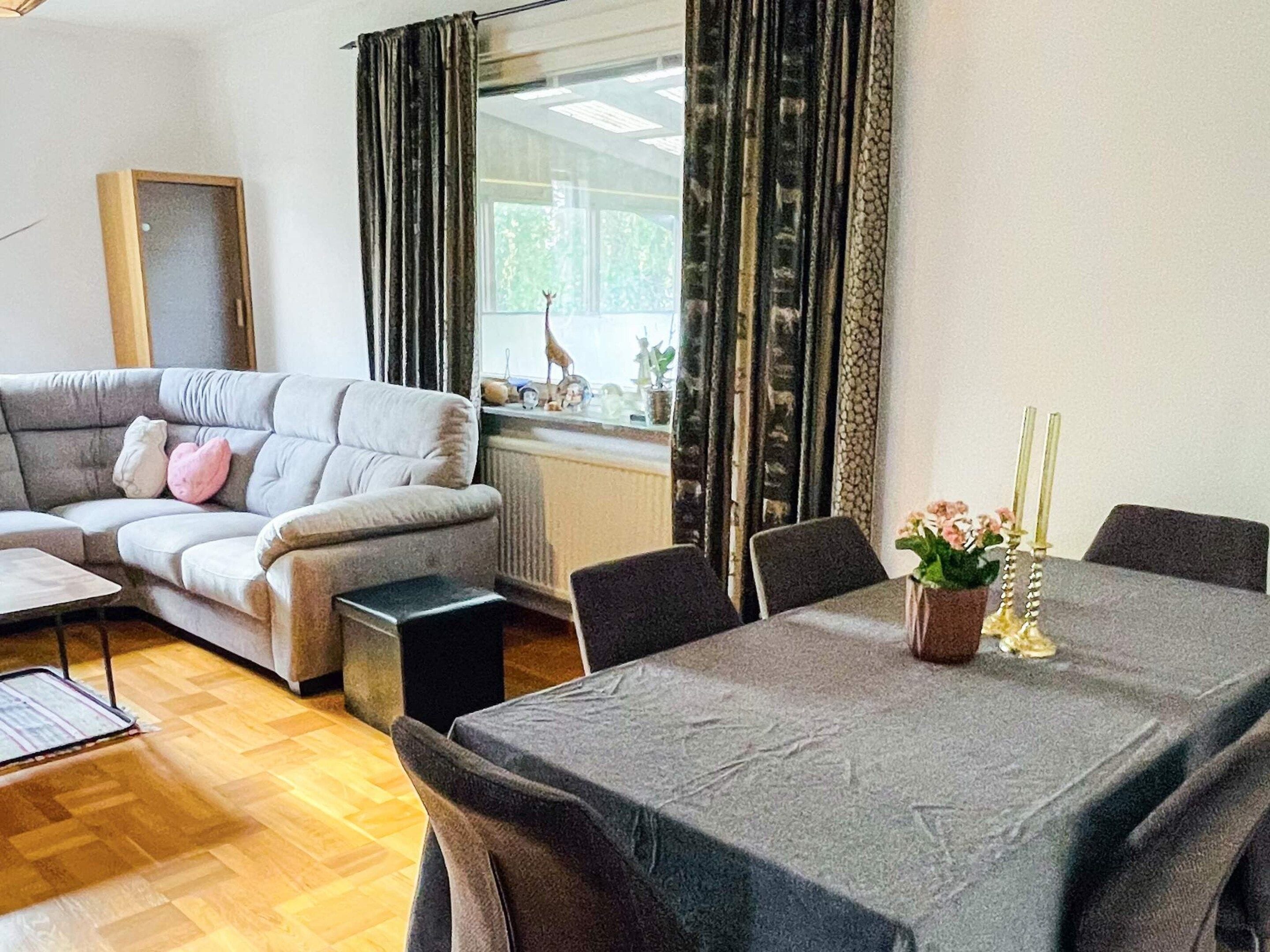 A living room with a grey sofa, a dining table with chairs, a small black speaker, and a window with curtains.