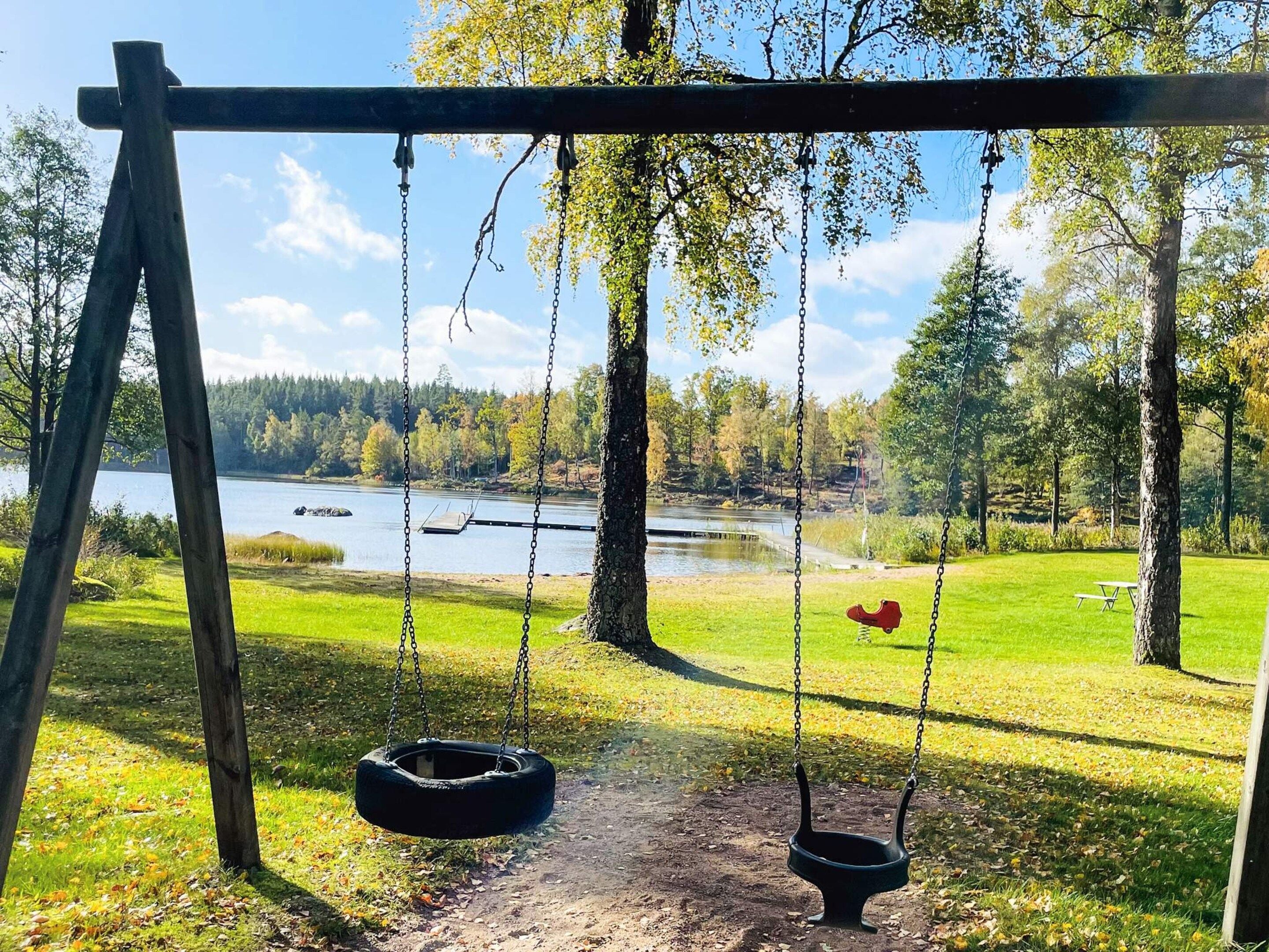 A wooden swing set with a tire swing and a bench, overlooking a lake with trees and a picnic table in a park.