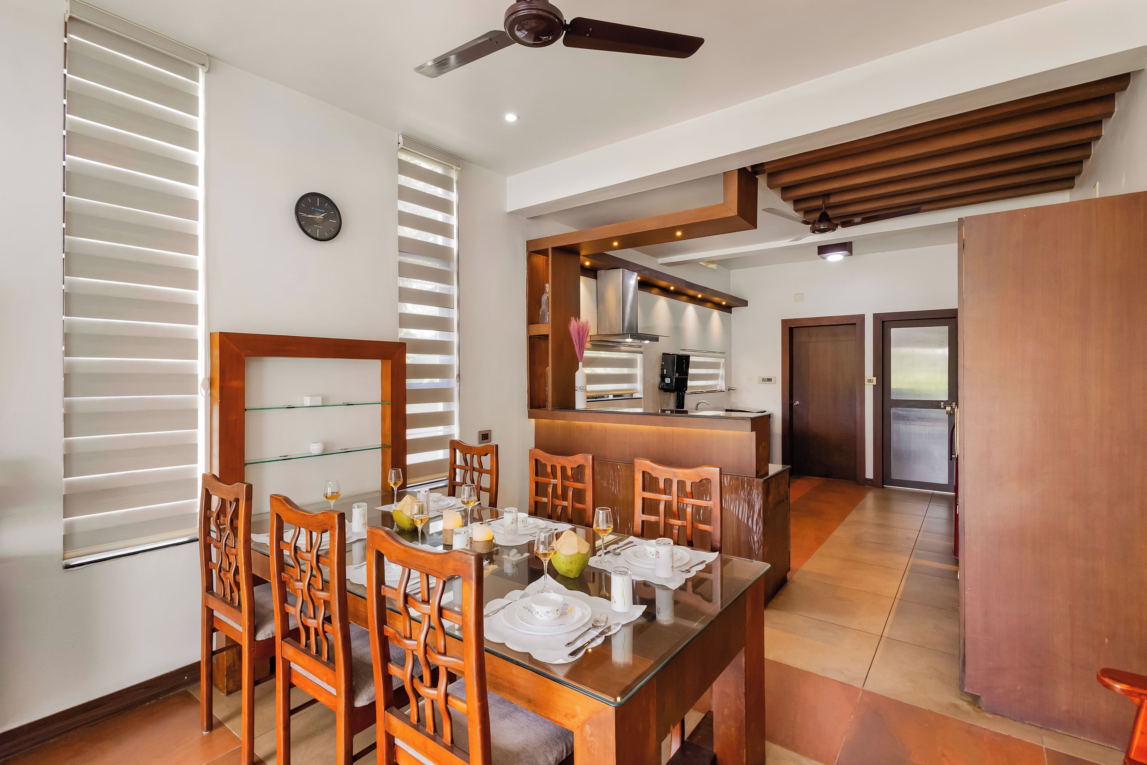A dining area with a glass table, wooden chairs, and a kitchen in the background.