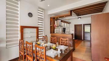 A dining area with a glass table, wooden chairs, and a kitchen in the background.