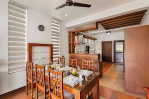 A dining area with a glass table, wooden chairs, and a kitchen in the background. - Glass River Retreat (Aluva)