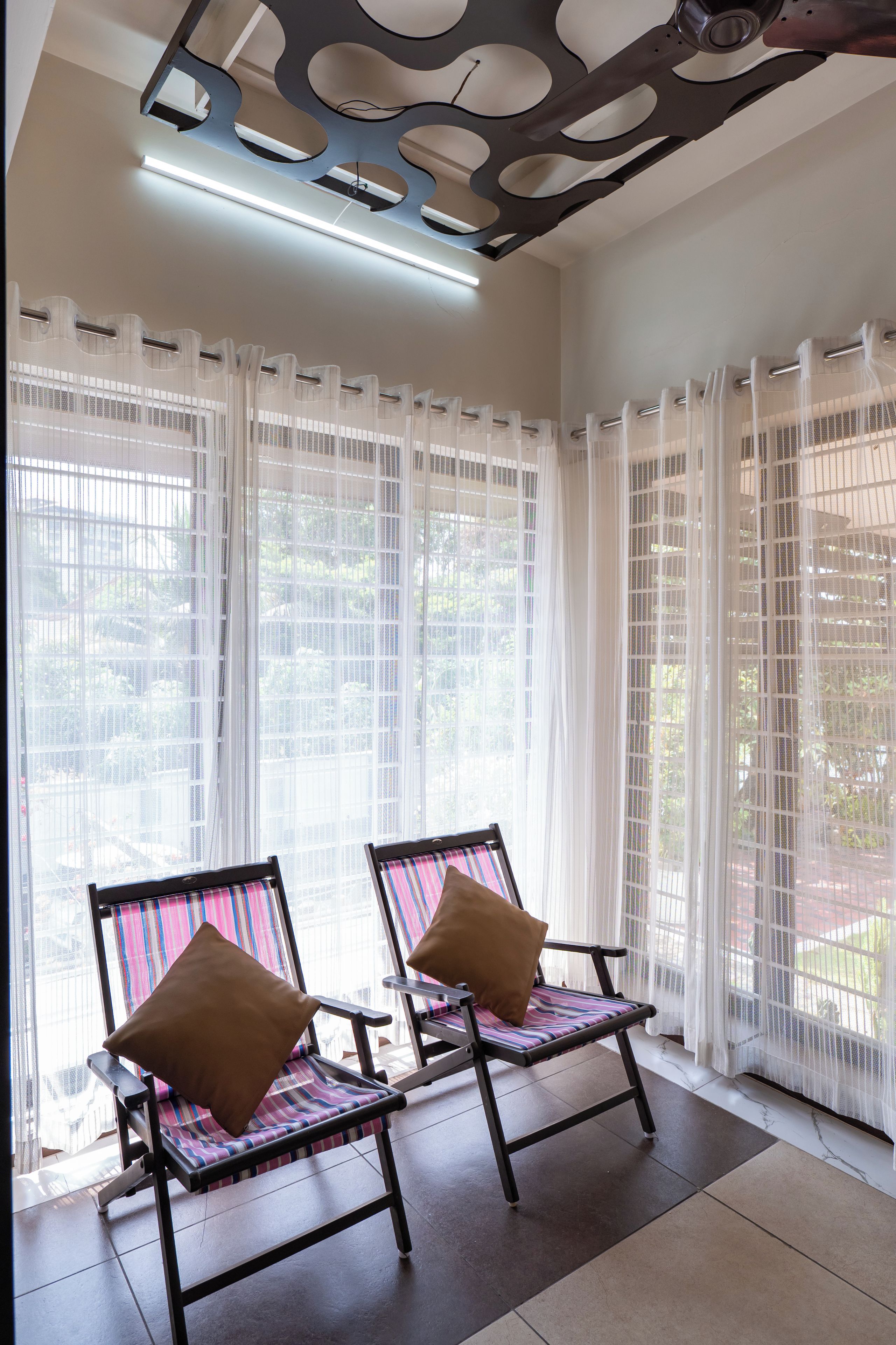 A room with two chairs, a ceiling with a unique design, and a view through large windows with sheer curtains.
