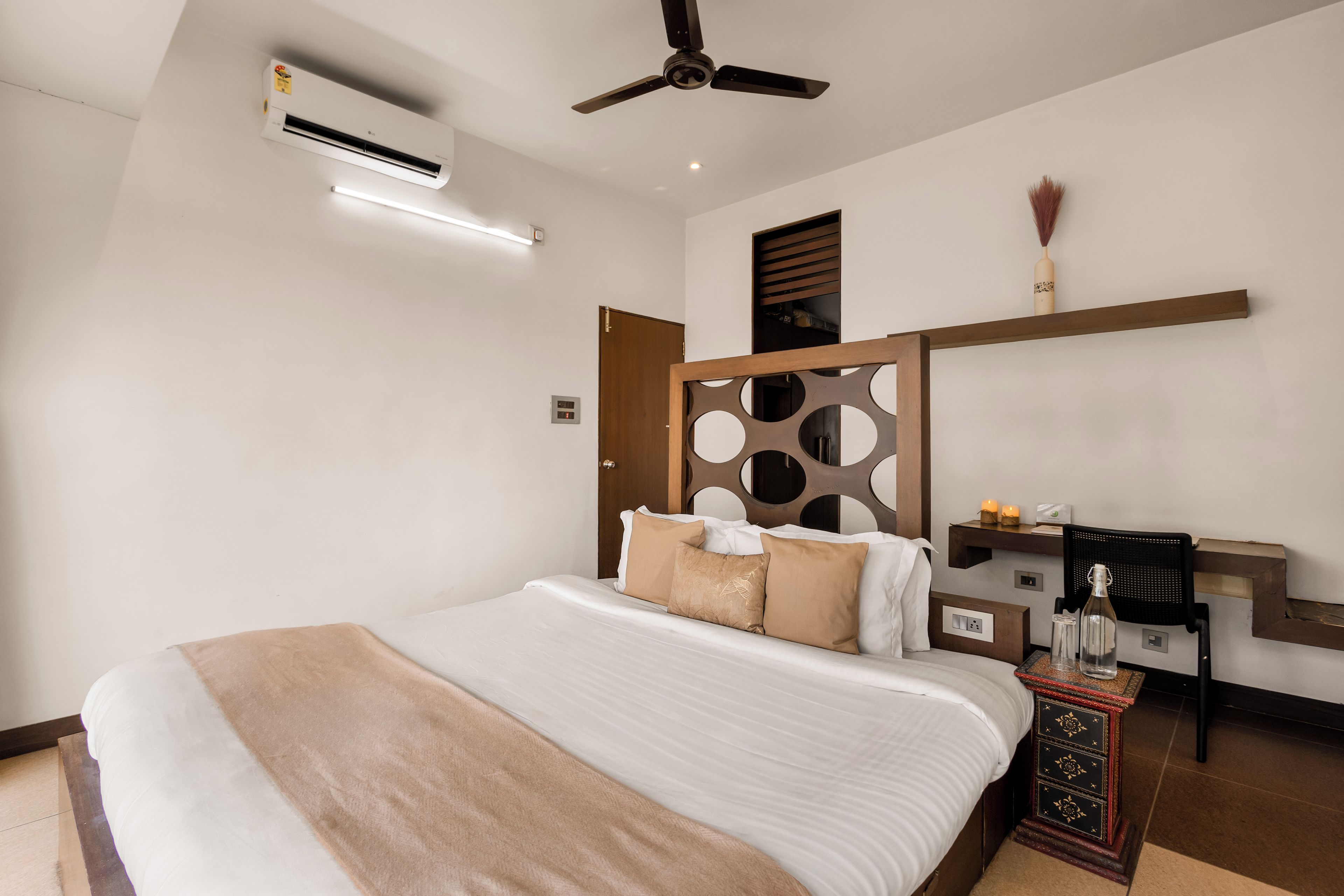 A modern hotel room with a large bed, a ceiling fan, an air conditioning unit, a bedside table with a lamp, and a decorative wall panel.