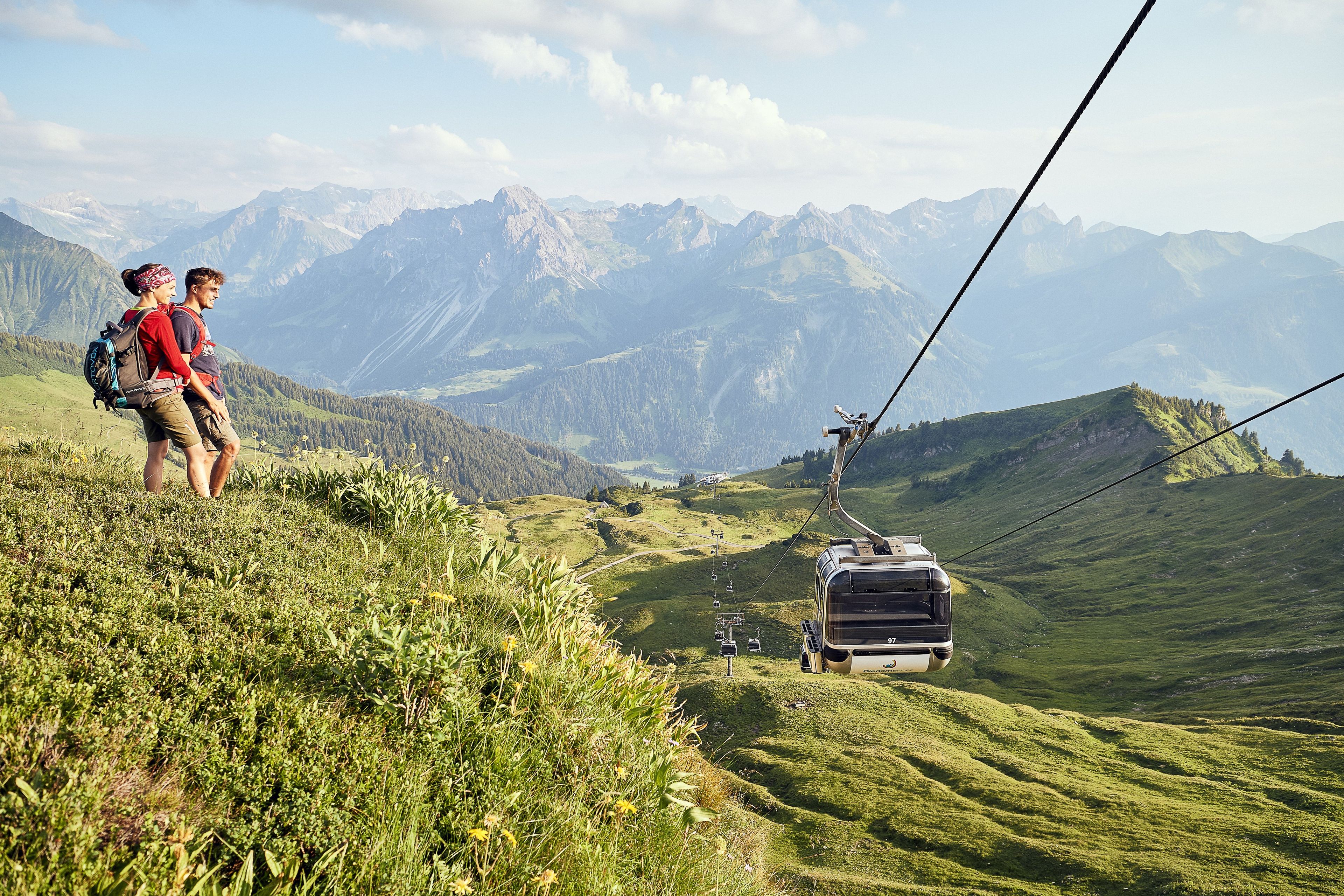 Un téléphérique se déplace au-dessus d’un paysage montagneux, avec des randonneurs sur une pente herbeuse.