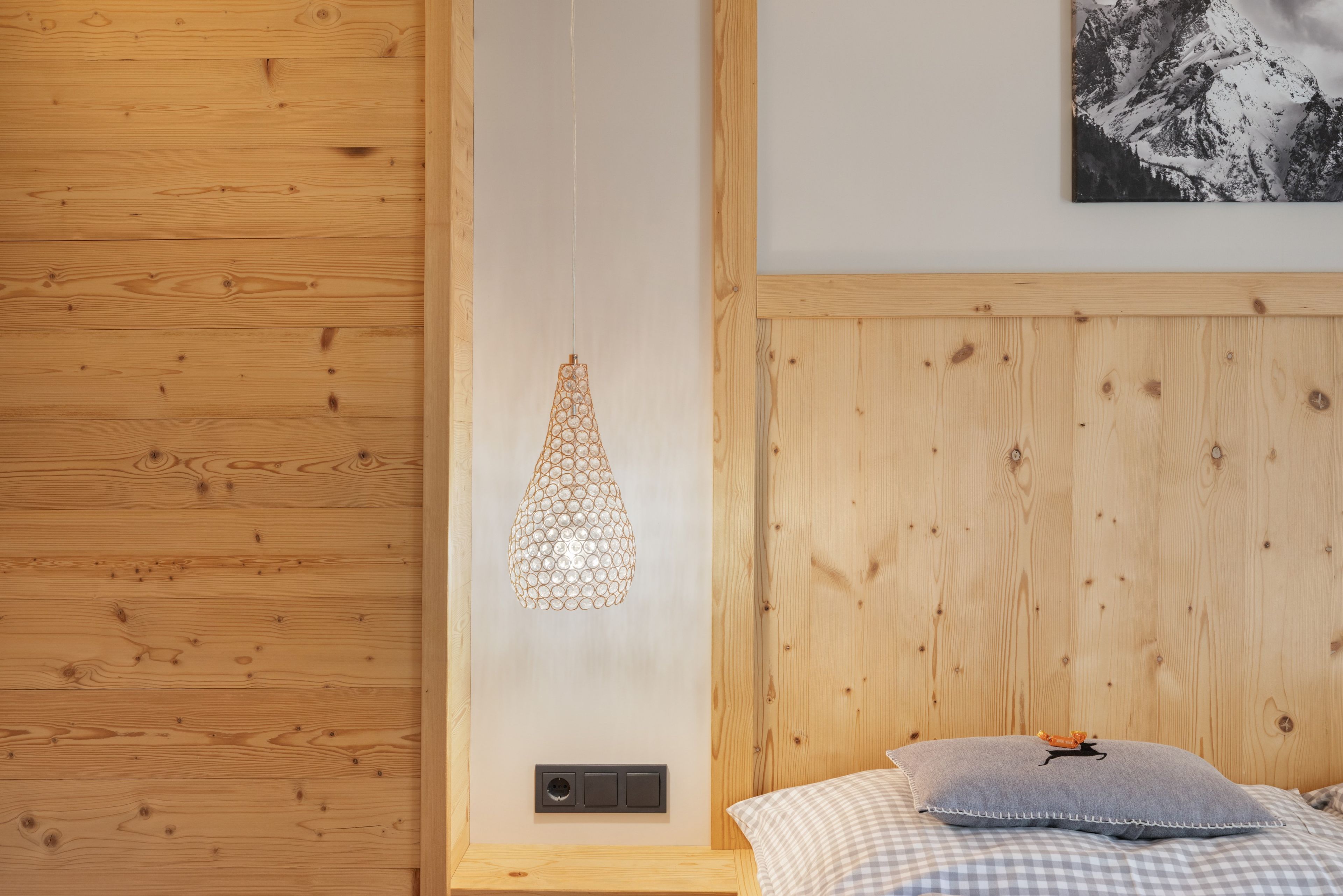 A bedroom with wooden paneling, a hanging light fixture, a bed with a checkered pillow, and a wall-mounted power outlet.