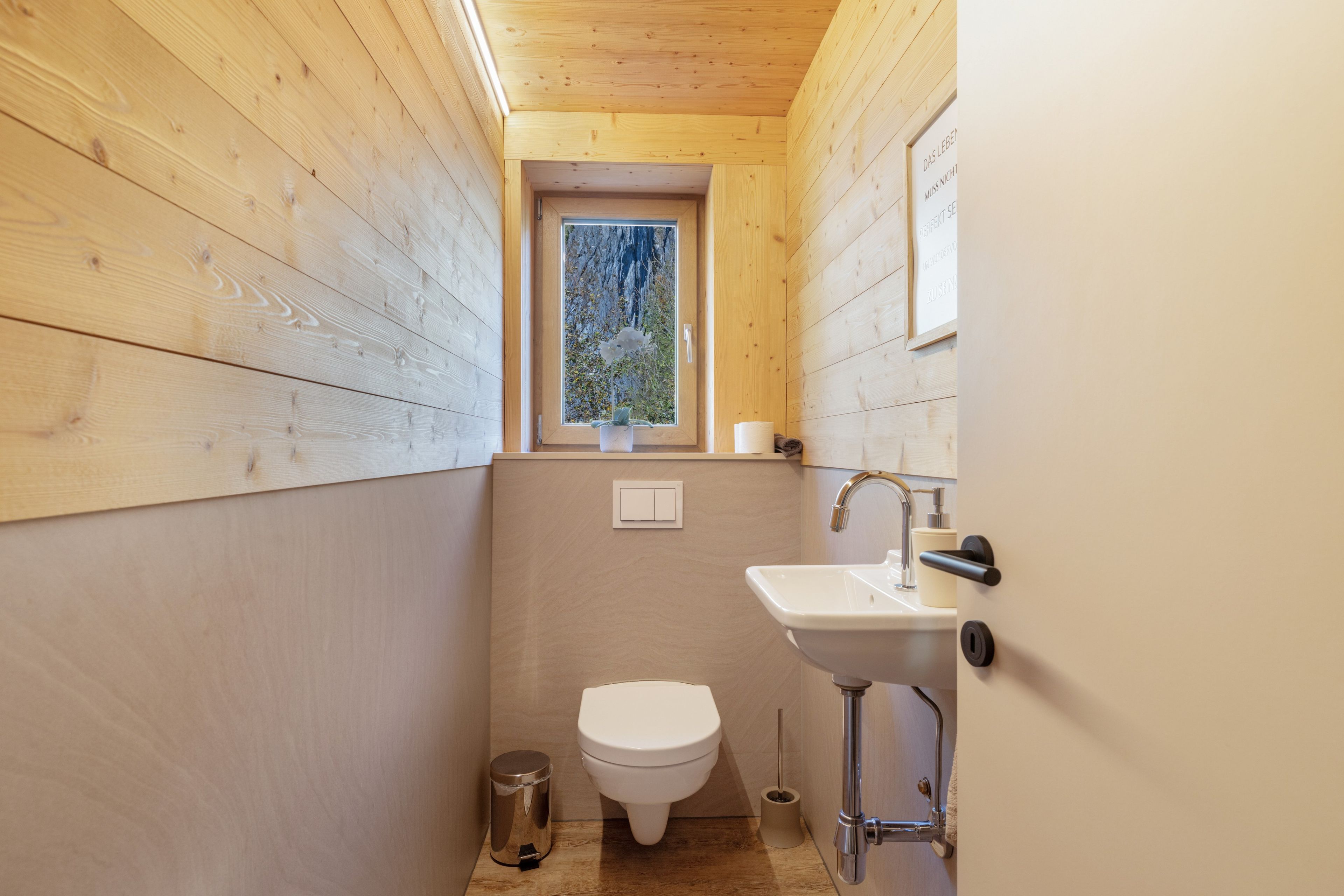 A compact bathroom with wooden walls, a white toilet, a sink, and a window with a view of trees.