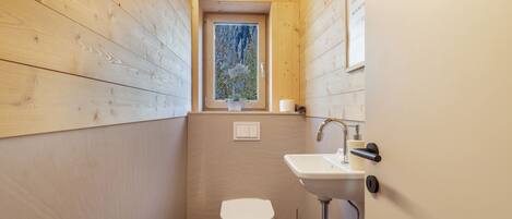 A compact bathroom with wooden walls, a white toilet, a sink, and a window with a view of trees.