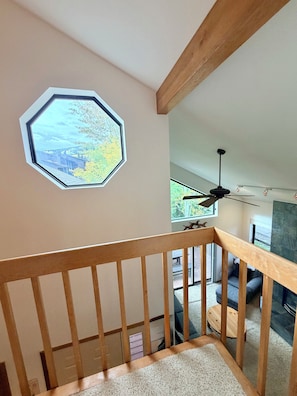 Eine Holztreppe führt zu einem Wohnbereich mit großem Fenster, Deckenventilator und Blick ins Freie.