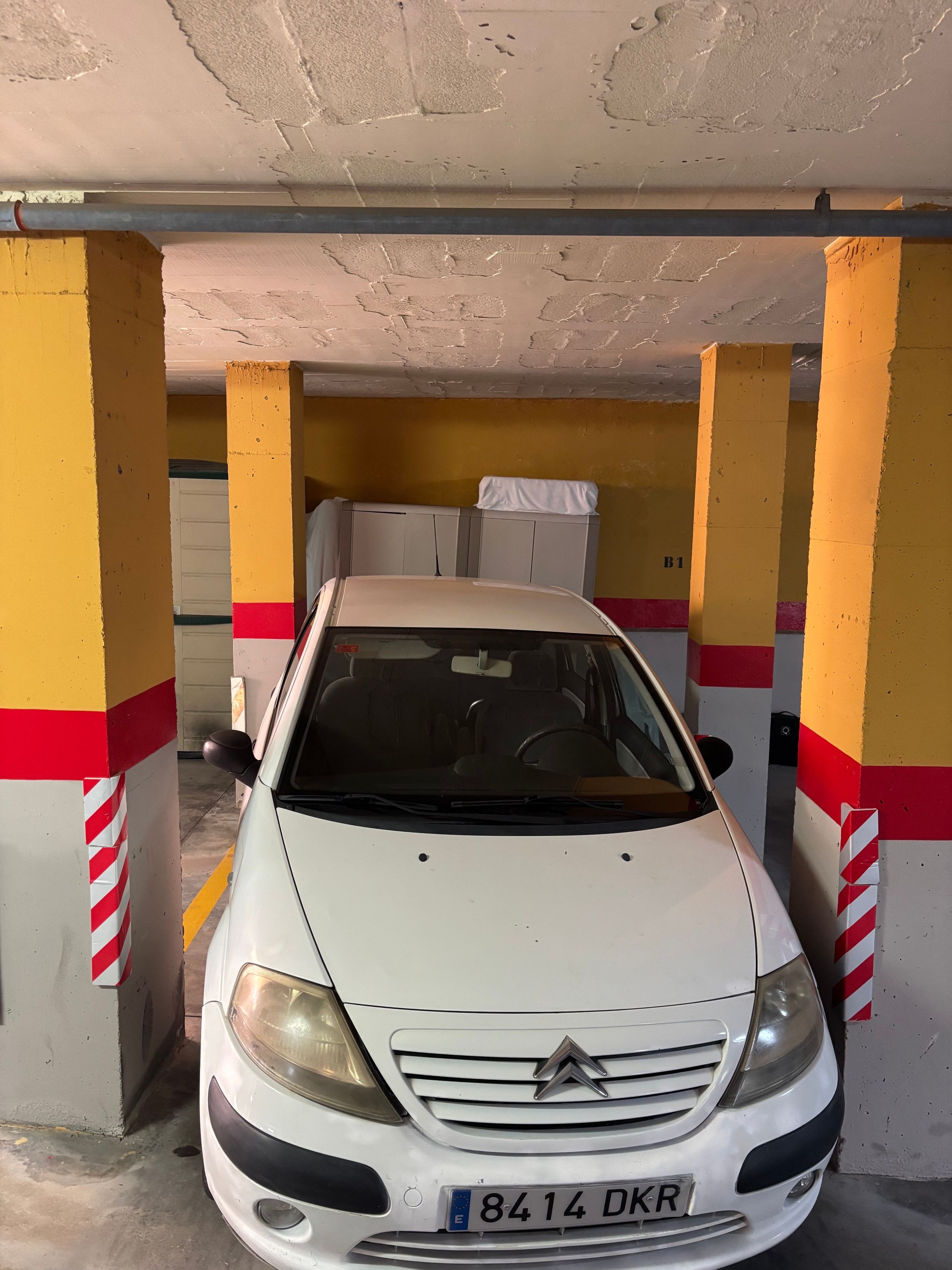 Ein weißes Auto steht in einer Parkgarage mit gelben und roten Säulen, einer Beton Decke und einer Tür im Hintergrund.