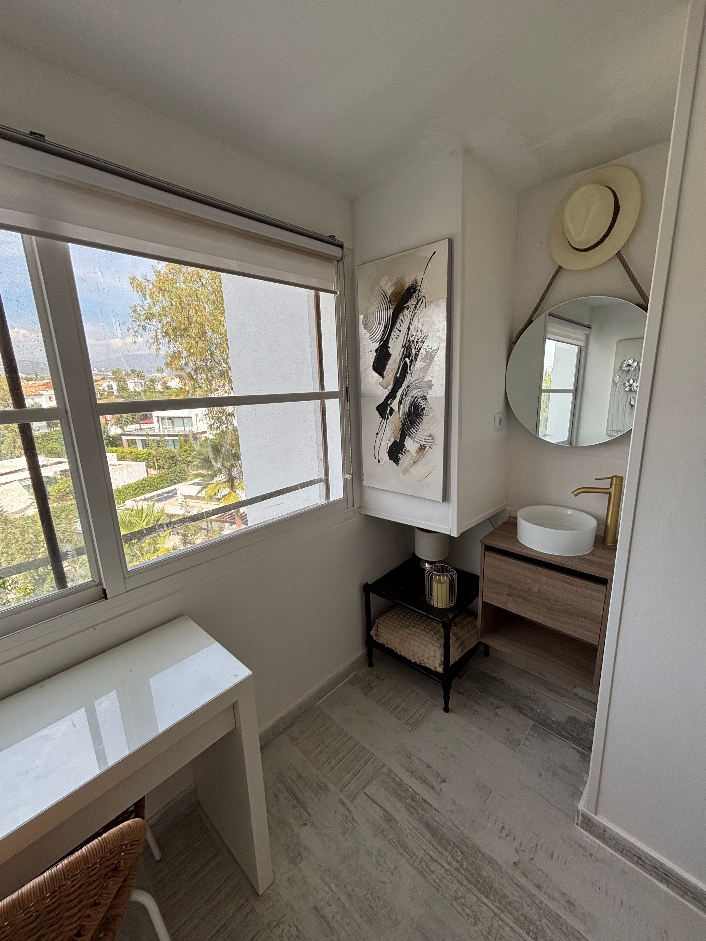 A bathroom with a white vanity, a round mirror, and a window with a view of buildings and trees.