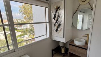 A bathroom with a white vanity, a round mirror, and a window with a view of buildings and trees.